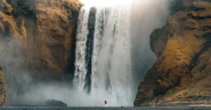 A stunning waterfall flows down a brown mountain with the sun shining above. A person in a red and white outfit stands in front of the waterfall.