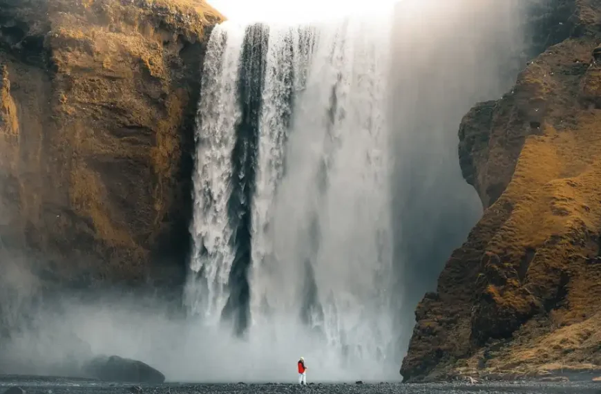 A stunning waterfall flows down a brown mountain with the sun shining above. A person in a red and white outfit stands in front of the waterfall.