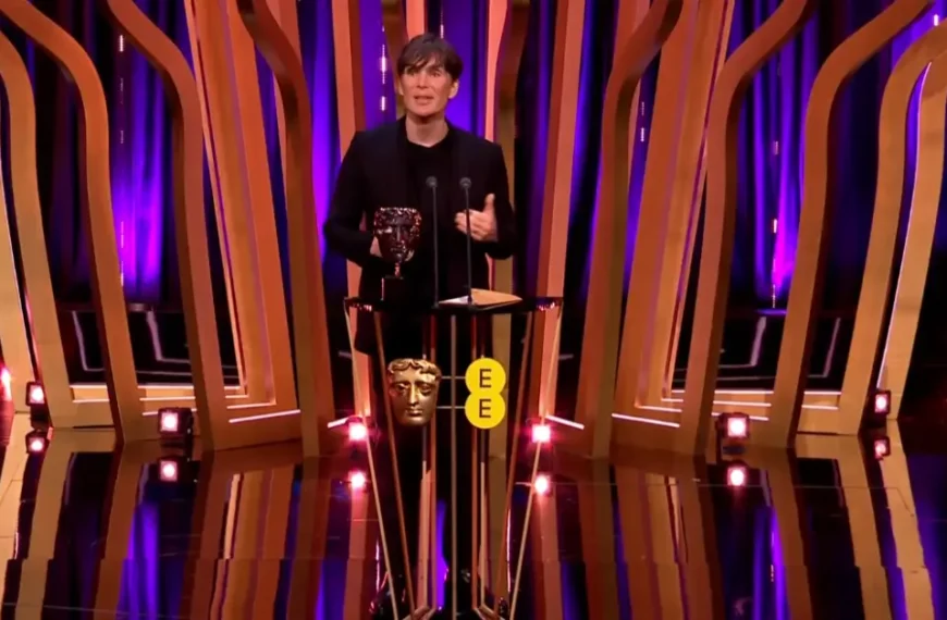 Cillian Murphy in a black suit, holding a Bafta Award, standing on a colorful stage, speaking into a microphone.