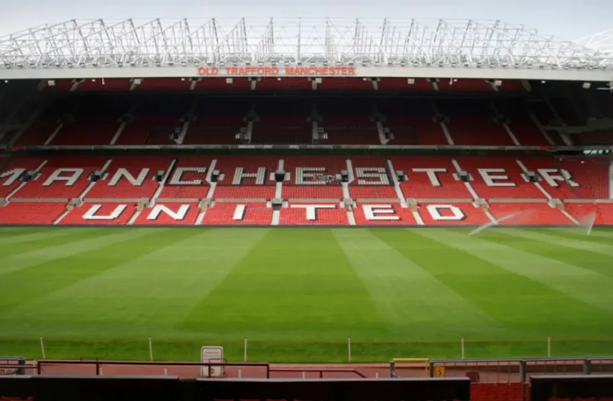 An empty football ground with red seats, featuring the visible name 'Old Trafford Manchester' and 'Manchester United' written on the chairs.