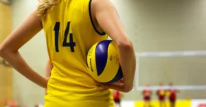 A determined female volleyball player in a yellow shirt with the number 14, holding a ball and standing on an indoor court with a blurred background.