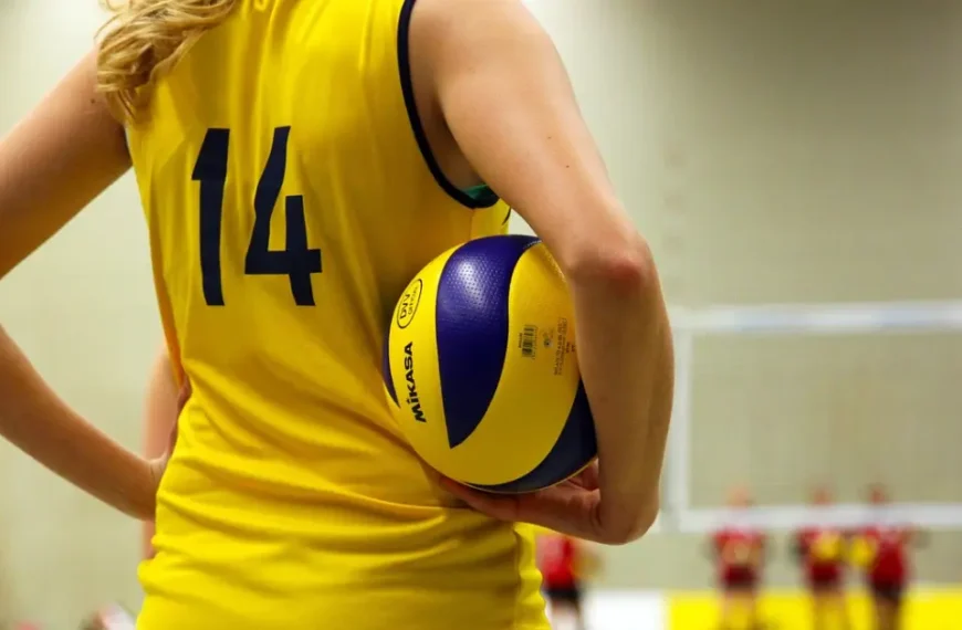 A determined female volleyball player in a yellow shirt with the number 14, holding a ball and standing on an indoor court with a blurred background.