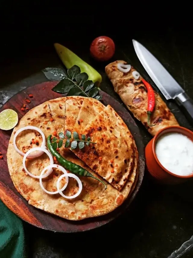 A sliced Aloo Paratha on a wooden plate, garnished with onion, green chili, and salt. A knife and tomato are seen beside it, along with a sour lemon.