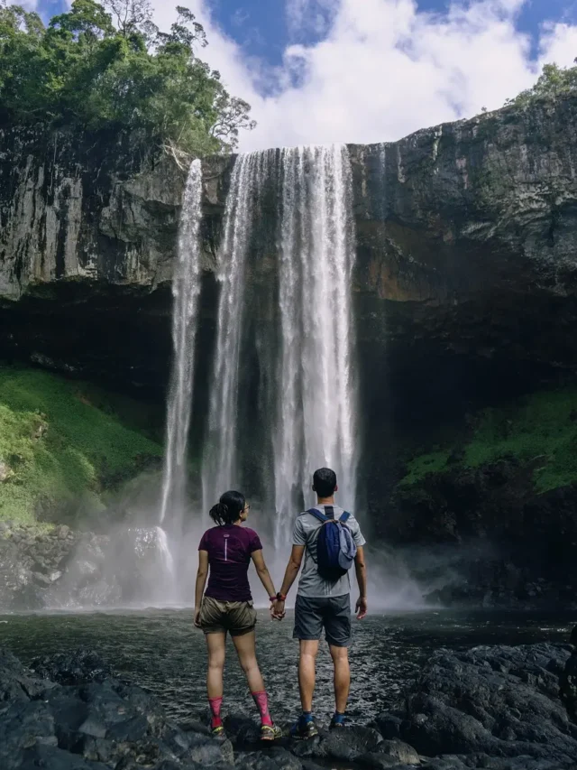 A couple, stands before a stunning waterfall, their hands clasped in unity. The picturesque backdrop of cascading water complements their adventurous spirit, creating a serene and romantic scene.